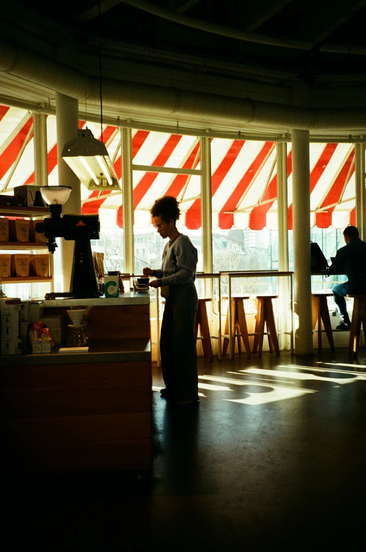 A barista prepares a drink in a Berlin café, with sunlight falling through striped awnings.